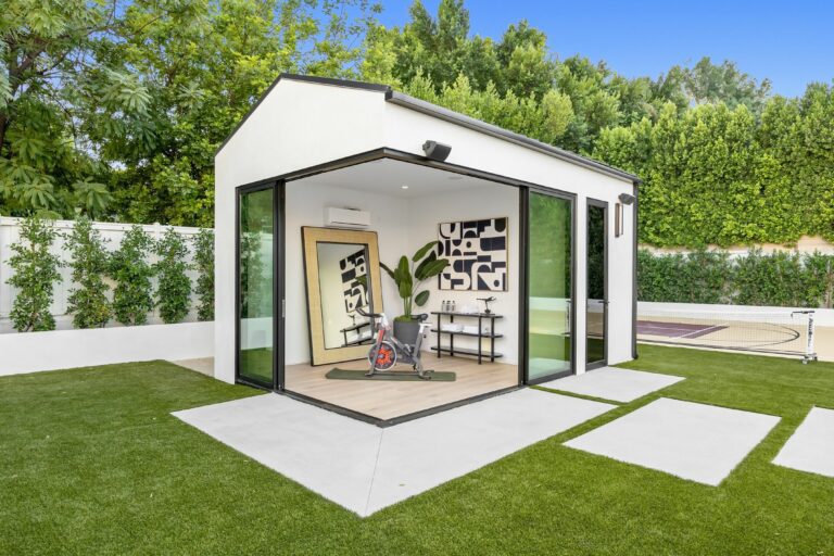A mid-century modern living room with designer furniture, an indoor garden with a fountain, large windows revealing a forest landscape at sunset, and an indoor potted tree.