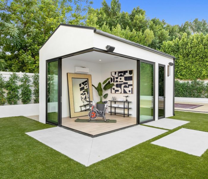 A mid-century modern San Jose, CA Home addition living room with designer furniture, an indoor garden with a fountain, large windows revealing a forest landscape at sunset, and an indoor potted tree.
