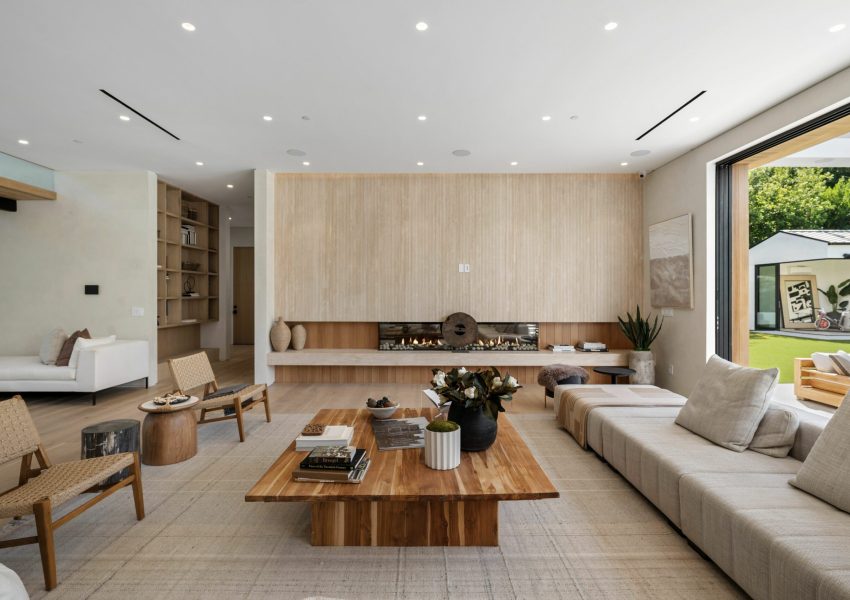 Alt text: A mid-century modern living room with designer furniture, an indoor garden with a fountain, large windows revealing a forest landscape at sunset, and an indoor potted tree.